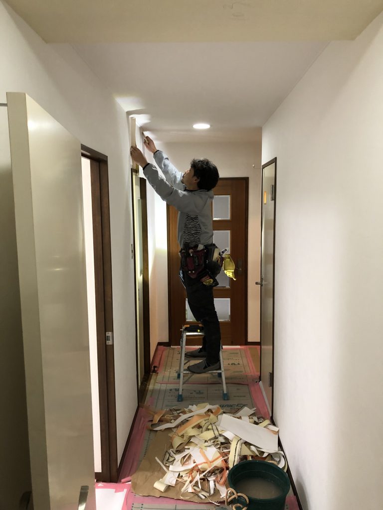 入間マンションリフォームで職人が廊下の壁のクロス張り替え作業をしている画像です。