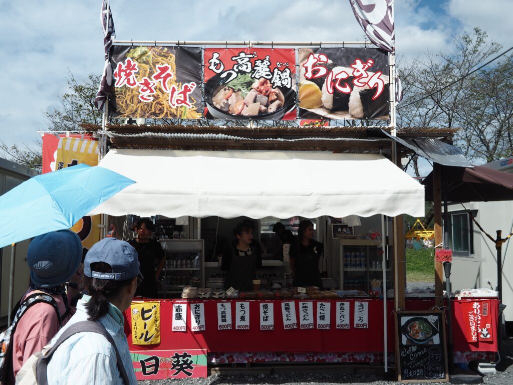 埼玉県日高市巾着田曼殊沙華公園の出店、おにぎりやたこ焼きや赤飯や焼きそばもつ鍋などが売っています。日傘をさした人や帽子をかぶった観光客と店員が3名写っています。