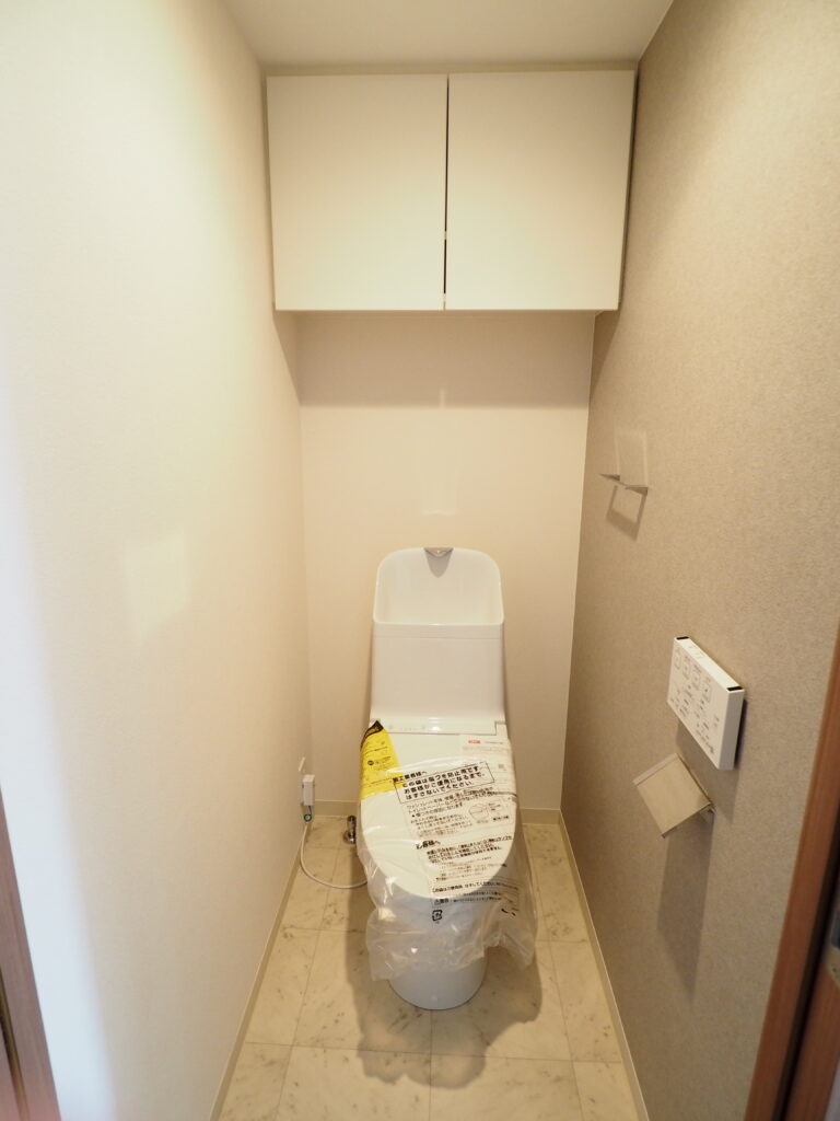 八王子市内中古マンションの施工後のトイレ画像、トイレの便器と背面収納、トイレットペーパーホルダー、ウォッシュレットリモコンが写っています。
