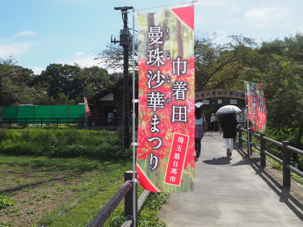 埼玉県日高市巾着田曼殊沙華公園の曼殊沙華まつりののぼりと橋を歩く人が写っています。