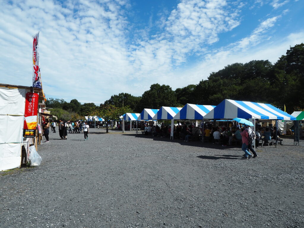 埼玉県日高市巾着田曼殊沙華公園の多目的スペース、テントの下にはベンチでくつろいでいる人がいます。出店が写っています。