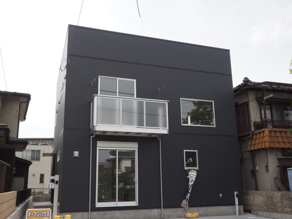 四角い形の戸建て住宅、外壁の色はとても濃いグレーで黒に近いです。窓が4つとベランダが写っています。 外用の水道があります。