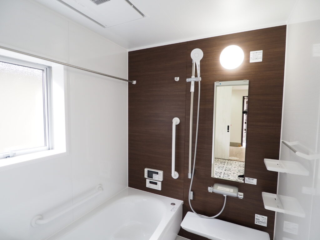 浴室の写真です。木目調のアクセントパネルとシャワー、鏡、浴槽、手摺、給湯器リモコンに浴室用テレビ、タオルハンガーに収納棚と照明が写っています。